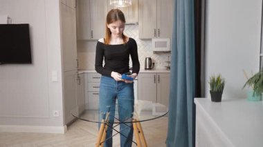 Genç kız titizlikle cam mutfak masasını mavi bezle siliyor, modern ev ortamında evin titiz bakımını ve temizliğini gösteriyor.