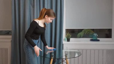 Genç kız mavi bezle cam masayı temizliyor, mavi perdelerle evi temizliyor ve pencere eşiğinde bitki yetiştiriyor.