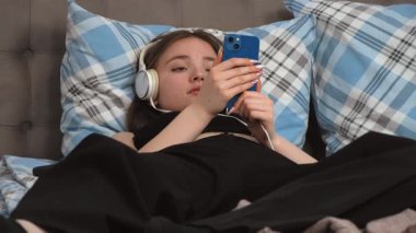 Kulaklıklı güzel bir genç kız, en sevdiği melodilere dalmış, gözleri kapalı yatakta yatarken akıllı telefondan müzik dinliyor.