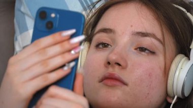 Kulaklıklı güzel bir genç kız, en sevdiği melodilere dalmış, gözleri kapalı yatakta yatarken akıllı telefondan müzik dinliyor.