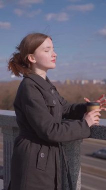 İçkiler ve insanların konsepti. Mutlu genç kadın ya da genç kız. Şehrin arka planında uzun saçlı, şık bir kızla kağıt bardaktan kahve içiyor.