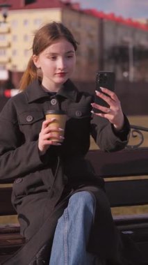 Genç bir kız kenarda oturmuş, elinde akıllı telefonuyla kahve içerken kentteki rahatlama anını seyrediyor ve selfie dikey video çekiyor.