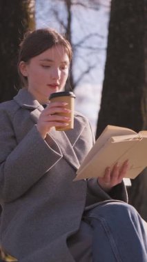 Sıcak güneş ışığı, kitap okuyan ve park ağaçlarının arasında sessizce oturup rahatça kahve içen genç bir kızı kucaklıyor. Dikey tefekkür videosu.