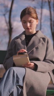 Sıcak güneş ışığı, kitap okuyan ve park ağaçlarının arasında sessizce oturup rahatça kahve içen genç bir kızı kucaklıyor. Dikey tefekkür videosu.