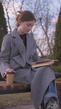 Gri bornozlu genç kız parkta kitap okuyor sıcak içkisini yudumlarken sakin bir sonbahar gününde sakin bir yalnızlık anı yaşıyor dikey videoda.