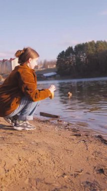 Gündelik giysiler giymiş genç bir kız su kenarında huzurlu bir anın tadını çıkarıyor, gölde yüzen ördekleri besliyor ve çömeliyor, doğanın barışçıl güzelliği ile çevrili.