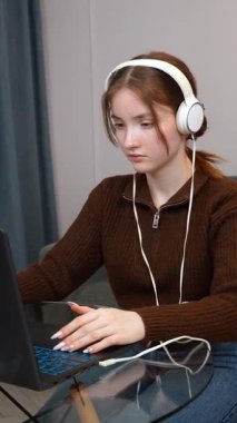 Kahverengi kazaklı ve kulaklıklı genç kız evden uzaktan çalışıyor, cam masada dizüstü bilgisayarla oturuyor, oyunlar oynuyor. dikey video