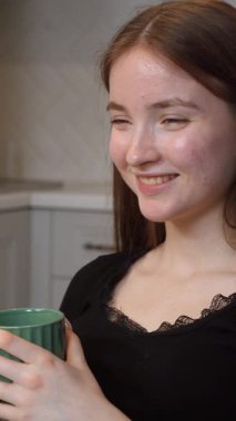 Genç bir kız elinde yeşil bir seramik bardak tutarken mutfaktaki sıcak havanın tadını çıkarırken huzurlu bir tatmin anı yaşıyor.