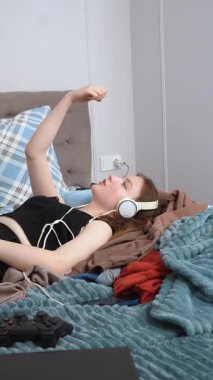 Rahatlamış genç bir kız yatağında dinleniyor, kulaklık takıyor, oyun seansından sonra müzikten keyif alıyor, evde dikey bir videoda huzurlu bir dinlenme anını kişiselleştiriyor.