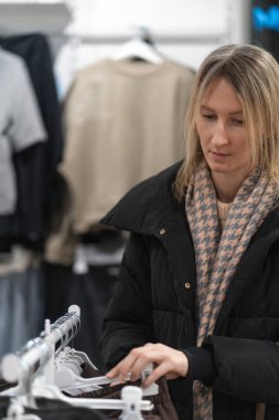 Şık kış kıyafetleri giymiş şık bir butikte askılardan elbise seçen ve gezen genç bir sarışın kadın içeride alışveriş deneyiminin tadını çıkarıyor. Dikey fotoğraf