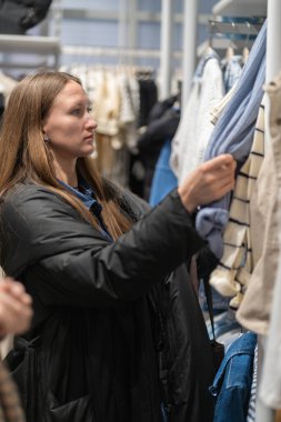 Kıyafet alışverişi yapan, moda mağazasında askıda asılı duran giysilere dokunan, perakende terapiden zevk alan ve satın alma kararı veren genç bir kadın. Dikey fotoğraf