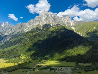Görkemli bir dağ zirvesinin büyüleyici panoramik görüntüsü açık mavi gökyüzünün altında parlak yeşil bir vadi üzerine uzun gölgeler bırakıyor Svaneti, Georgia 'da tüylü bulutlar var.