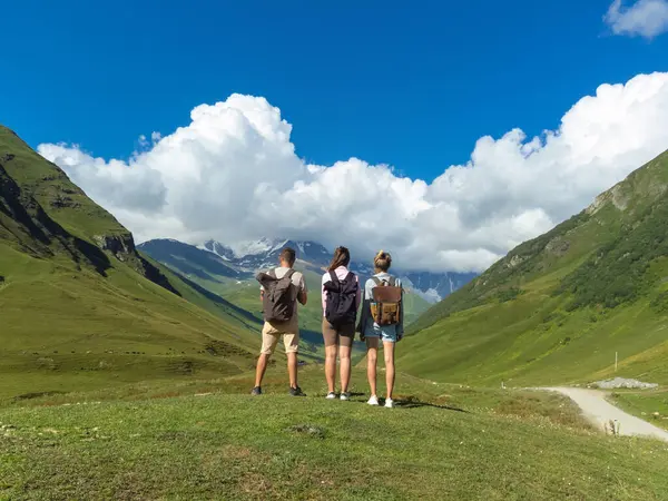 Sırt çantalı üç turist, yürüyüş ve yürüyüş maceraları için popüler bir yer olan Gürcistan 'ın svaneti bölgesindeki yeşil vadi ve kar kaplı zirvelerin nefes kesici manzarasının keyfini çıkarıyorlar.