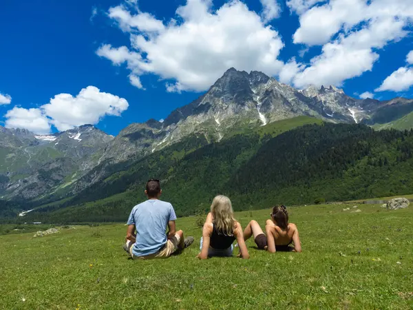 Üç turist çimenli bir çayırda soluk soluğa kalmış, parlak mavi bulutlu svaneti dağının altında Tetnuldi Dağı 'nın görkemli manzarasında dinleniyor.