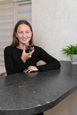 Gülümseyen genç bir kadın akıllı telefonuyla sesli bir mesaj kaydederken modern dairesinde mutfak tezgahına yaslanmış, rahat bir şekilde bağlantının keyfini çıkarıyordu..
