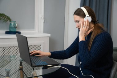 Kulaklık takan genç bir kadın evde dizüstü bilgisayarla çalışıyor, video konferansına katılıyor ya da işini yönetirken müzik dinliyor.
