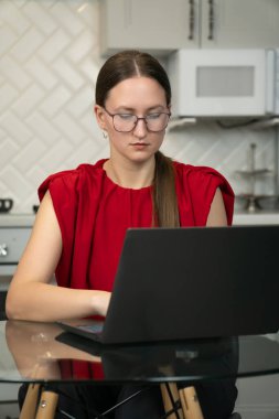 Gözlüklü ve kırmızı tişörtlü genç bir kadın, modern bir mutfakta dizüstü bilgisayarıyla çalışmaya odaklanıyor ve bu da evden çalışma tarzını gösteriyor. Dikey fotoğraf