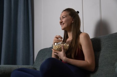 Gülümseyen genç kadın rahat bir koltukta oturup elinde cam kaseyle evde ilginç bir film izlerken patlamış mısır yemekten zevk alıyor.