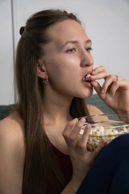 Kanepede rahat bir şekilde oturan genç bir kadın film izlerken cam bir kaseden patlamış mısırın tadını çıkarıyor evde huzurlu ve rahatlatıcı bir akşamın tadını çıkarıyor. Dikey fotoğraf