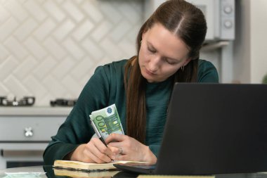 Kadın dikkatli bir şekilde ev ekonomisini yönetiyor. Titizlikle bir deftere harcamalarını yazıyor. Avro banknotlarını tutuyor ve mutfaktaki dizüstü bilgisayarını kullanıyor.