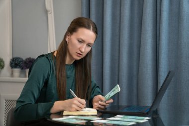 Kişisel hesaplarını yöneten, Avro banknotları ve bir not defteri kullanarak gelir ve giderlerini hesaplayan genç bir kadın.
