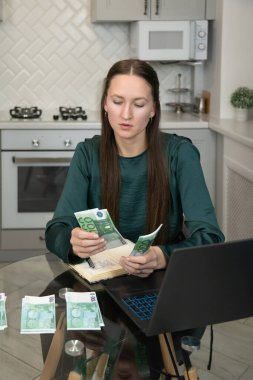 Mutfak masasındaki avro banknotlarını titizlikle sayarken ev ekonomisini yöneten genç bir kadın, online bankacılık ve bütçe işleri için dizüstü bilgisayar kullanıyor. Dikey fotoğraf