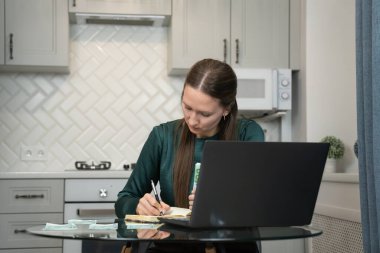 Bilgisayarı kullanarak ve mutfak masasında euro banknotlarını sayarak ev giderlerini hesaplarken, ev bütçesini ve kişisel finansmanı yöneten odaklı kadın defterine notlar yazıyor.