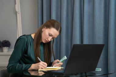 Avro banknotlarını sayarken ve not defterine notlar yazarken, ev bilgisayarında rahat bir ofis ortamında çalışan genç bir kadın.