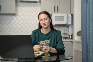 Bize dolar banknotları sayarak ev bütçesini yöneten, mutfaktaki cam masada oturan, dizüstü bilgisayarda çalışan ve not defterine notlar yazan genç bir kadın.