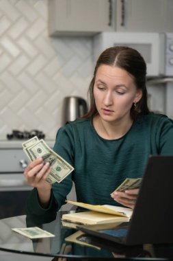 Ev ekonomisini yöneten genç bir kadın. Mutfak masasında dizüstü bilgisayar ve defterle oturup finansal sorumluluk ve ev bütçesini gösterirken bize dolar banknotlarını dikkatlice sayıyor.