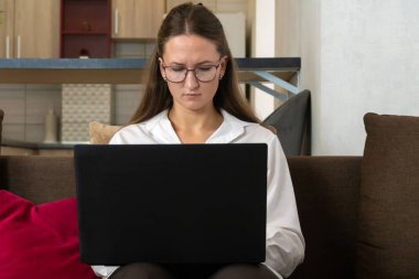 Gözlüklü ve beyaz tişörtlü genç bir kadın şık bir ev ofisteki koltukta rahat bir şekilde oturuyor uzaktan çalışma ve internet iletişimiyle uğraşırken dizüstü bilgisayarına odaklanıyor.