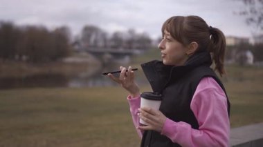 Açık havada, kahverengi saçlı, pembe kazaklı, siyah yelekli, elinde bir fincan kahve tutan ve akıllı telefonuna sesli mesaj gönderen genç bir kadın.