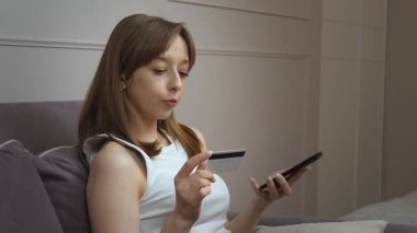 Oturma odasındaki kanepede rahatça dinlenen, akıllı telefondan online alışveriş yapan, kredi kartını kullanarak güvenli bir şekilde ödeme detaylarını giren bir kadın.