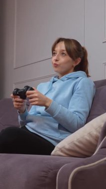Video oyunları oynayan, elinde kontrolörle zaferi kutlayan, oturma odasında kanepede oturan, oyun seansını kazanmanın sevincini ve heyecanını dile getiren genç bir kadın.