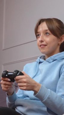 Video oyunları oynayan, elinde kontrolörle zaferi kutlayan, oturma odasında kanepede oturan, oyun seansını kazanmanın sevincini ve heyecanını dile getiren genç bir kadın.