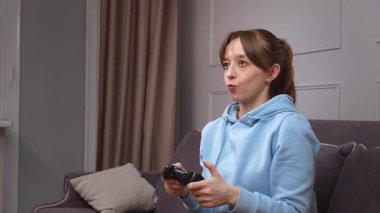 Kumanda panelinde joystick ile video oyunu oynayan tutkulu genç bir kadın, rahat bir şekilde kanepede oturuyor ve evde boş zamanlarında dijital eğlenceye dalıyor.