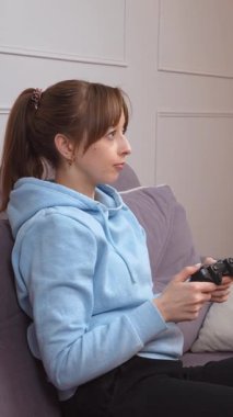 Günlük mavi kapüşonlu genç kadın video oyunlarından hoşlanıyor, kablosuz kontrolörle yoğun bir şekilde etkileşimde, oyun sırasında dinamik duygu aralığını konsol dikey video oturumunda gösteriyor