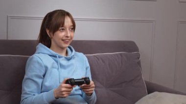 Günlük mavi kapüşonlu genç kadın video oyunlarından hoşlanıyor, kablosuz kontrolörle yoğun bir şekilde etkileşimde, oyun oturumunda dinamik duygu aralığı gösteriyor.
