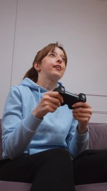 Günlük mavi kapüşonlu genç kadın video oyunlarından hoşlanıyor, kablosuz kontrolörle yoğun bir şekilde etkileşimde, oyun sırasında dinamik duygu aralığını konsol dikey video oturumunda gösteriyor