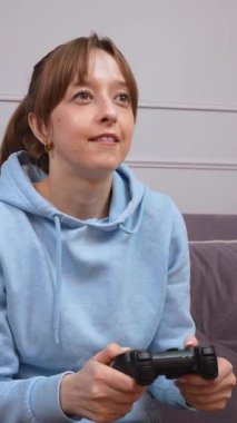 Günlük mavi kapüşonlu genç kadın video oyunlarından hoşlanıyor, kablosuz kontrolörle yoğun bir şekilde etkileşimde, oyun sırasında dinamik duygu aralığını konsol dikey video oturumunda gösteriyor