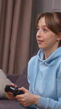 Günlük kıyafetlerle video oyunu oynayan genç bir kadın video oyunlarında kumanda panelinde, oturma odasında kanepede, heyecan verici bir oyun ve dikey video oyunu.