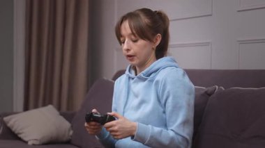 Günlük kıyafetlerle video oyunu oynayan genç bir kadın video oyunlarında kumanda panelinde, kanepede oturma odasında heyecanlı bir oyun oynuyor.