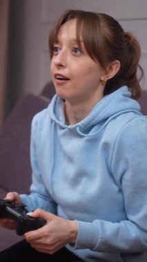 Günlük kıyafetlerle video oyunu oynayan genç bir kadın video oyunlarında kumanda panelinde, oturma odasında kanepede, heyecan verici bir oyun ve dikey video oyunu.