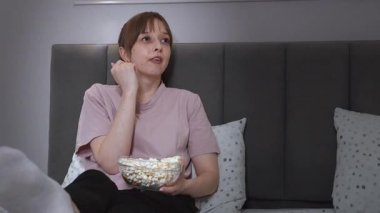 Yumuşak yastıklarla çevrili, rahat bir şekilde yatakta oturan bir kadın, evde rahat bir akşam boyunca gevşemiş film izlerken patlamış mısırın tadını çıkartıyor.