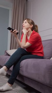 Oturma odasındaki koltukta rahat bir şekilde oturan rahat bir kadın elinde uzaktan kumandayla televizyon kanallarını değiştirirken evde dikey video izlemenin keyfini çıkarıyor.