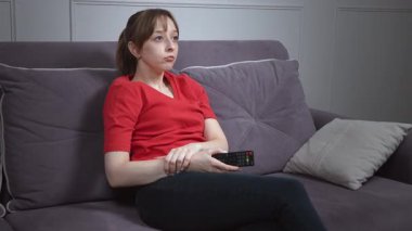 Kanepede dinlenen, kırmızı tişört giyerken televizyon kanallarını değiştiren, rahat bir konum bulan ve evde boş zamanlarında huzurlu bir anın tadını çıkaran bir kadın.