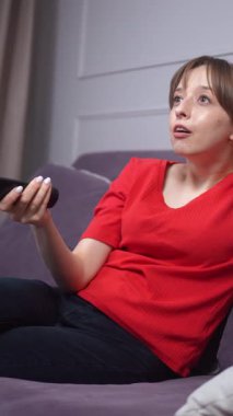 Kırmızı tişörtlü genç bir kadın rahat gri bir kanepeye oturur, televizyon izler, uzaktan kumandayla kanal değiştirir, ve elleri başının arkasında dikey video ile dinlenir.