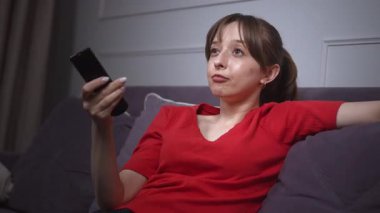 Kırmızı gömlekli genç bir kadın, elinde uzaktan kumandayla koltukta oturuyor ve televizyon seyrediyor. Kanallarda dolaşırken yüz ifadesi değişiyor.