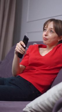 Kırmızı gömlekli genç bir kadın, elinde uzaktan kumandayla koltukta oturuyor ve televizyon seyrediyor. Kanalları dikey gezerken yüz ifadesi değişiyor.