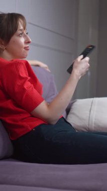 Kadın rahat bir şekilde yumuşak koltukta oturuyor, rahat bir şekilde televizyon kanallarını değiştiriyor. Uzaktan kumandayla rahat bir şekilde oturma odasında dikey video seyrediyor.